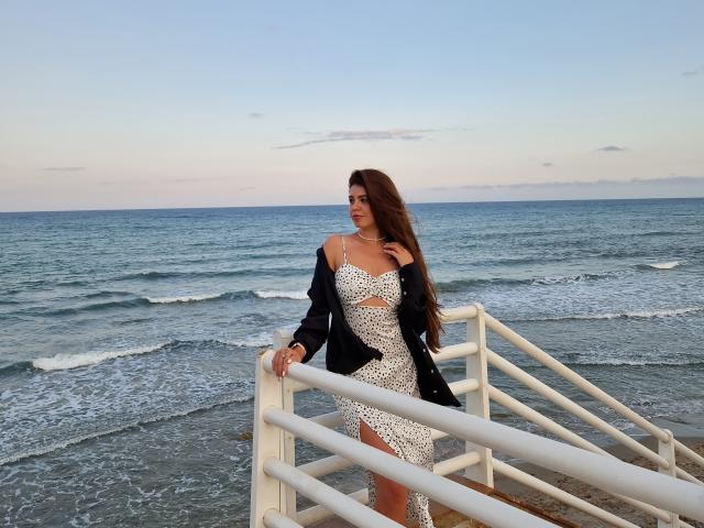 ValeriMair stands on a beach staircase, wearing a white, leopard-print dress with cutouts, black jacket, and long brown hair. Ocean waves in the background.