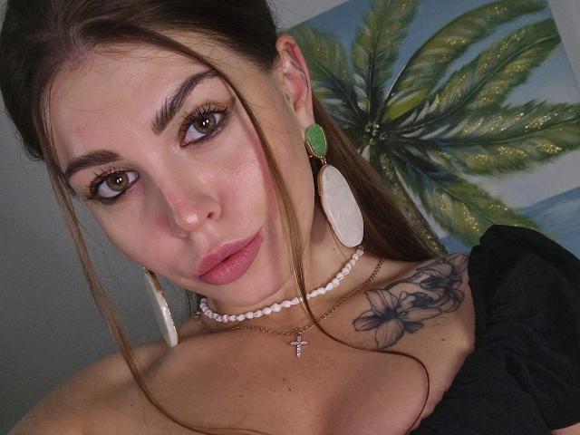 ValeriMair has brown eyes, pink lips, and a tattoo on her right shoulder. She's wearing green earrings, white necklace, and black top, showing her bare boobs.