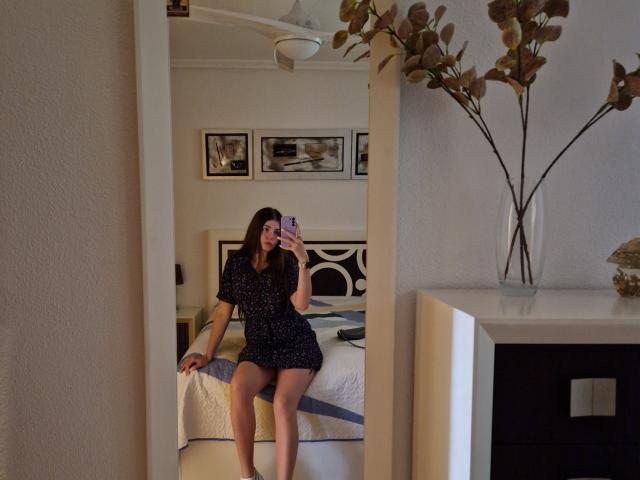 ValeriMair, sitting on a bed, wearing a black, short-sleeved, polka-dot dress, showing her ass, holding a phone, in front of a mirror. Bedroom with white walls.