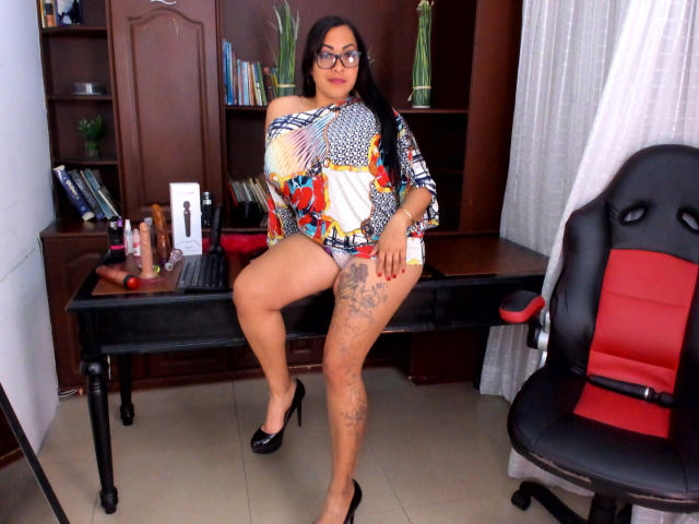 MellissaCortez with glasses, tan skin, and long dark hair, wearing a colorful off-shoulder top, black high heels, and no pants, showing her tattooed thigh and big ass.