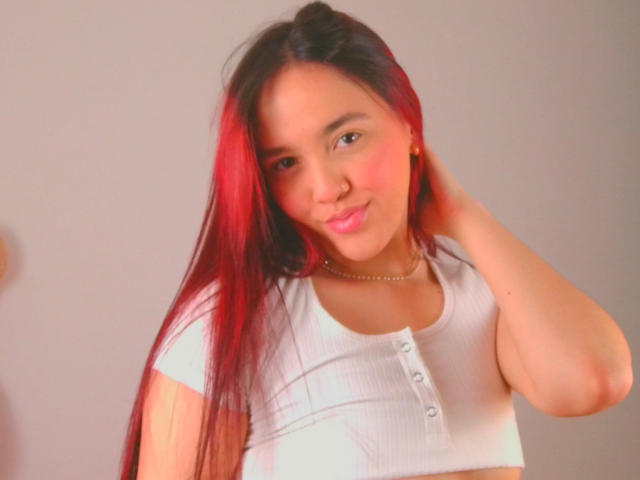 Milliee, con cabello largo de color rojo y negro, vistiendo una camiseta blanca de corte corto con botones, labios rosados y un collar de cadena de oro, tocándose la cabeza..