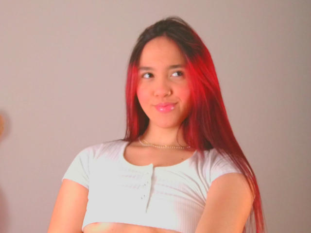 Milliee with long red-highlighted hair, white crop top, small tits, subtle smile, pink lips, light skin, gold necklace, looking to the side, soft red lighting on her face.