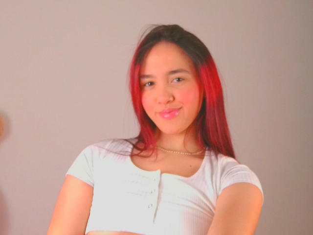Milliee with red and black hair, wearing a white shirt, has her arms crossed, smiling slightly, wearing a gold necklace. She has a cute face and pink blush.