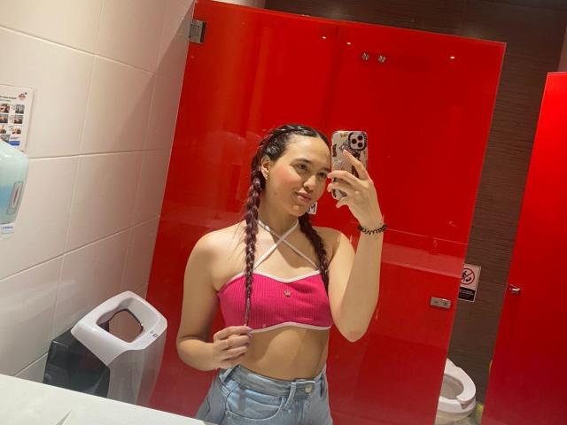 Milliee with pink bralette, exposing her small tits, and blue jeans, taking a mirror selfie in a red bathroom, holding her phone with braid-covered hands.