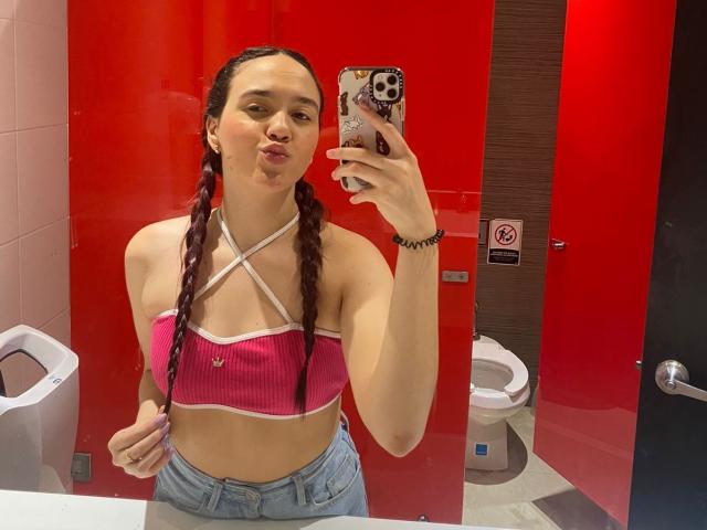 Milliee mit Zöpfen, rosa geripptem Crop-Top, gekreuzten Trägern, die Brüste zeigen, geschürzten Lippen, hält ein Handy in einem roten Badezimmer, trägt hoch taillierte Jeans, ein schwarzes Armband..