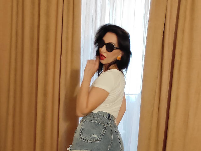 SimoniGrace, avec ses cheveux foncés, son rouge à lèvres rouge, ses lunettes de soleil noires, son haut blanc et son jean bleu déchiré, se tient devant des rideaux beige, touchant son menton, et montrant ses fesses rondes..