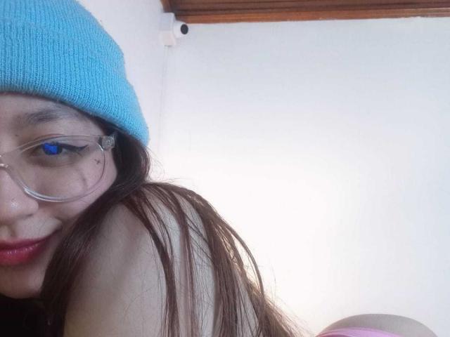 Alissa Brown met rood haar, een zwarte bandeau, een choker, die een spijkerbroek vasthoudt, met een tatoeage aan de linkerkant, die naar de camera kijkt, een blauw-witte geruite muur, neonlichten, een armbandje om de rechterpols..