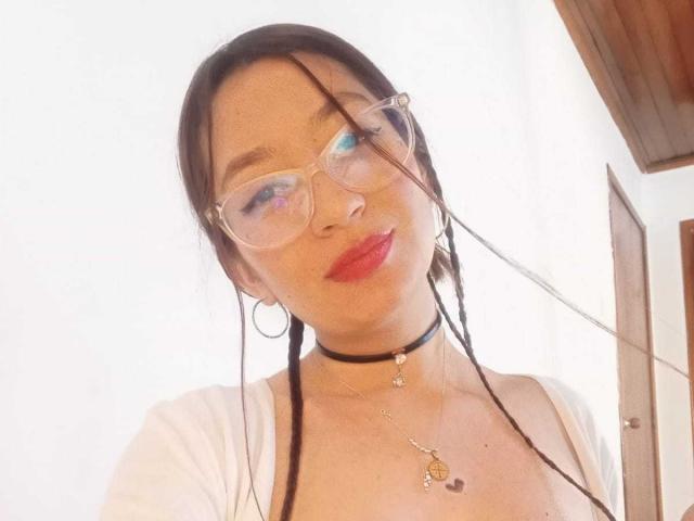 VioletaBlair con lunghi capelli castani, un asciugamano rosso che le copre il seno, una collana girocollo, sorridente, soffitto in legno sopra, che tiene l'asciugamano con la mano sinistra, angolazione leggermente bassa..