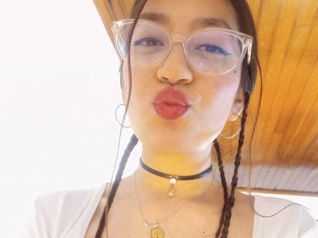VioletaBlair, con cabello castaño largo, brazos levantados, sonriendo, con lápiz labial rojo, sin top, mostrando pequeños senos, collar negro, techo de madera, pintura abstracta colorida en el fondo..