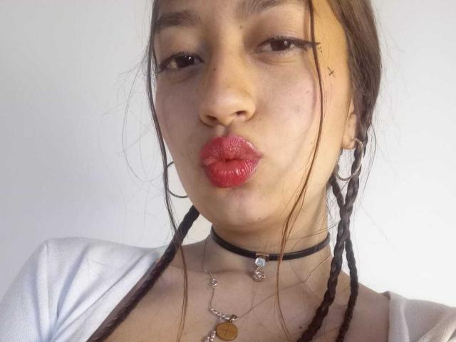 VioletaBlair, avec des lunettes, du rouge à lèvres rouge et des tresses, souriante, portant un collier noir avec une petite gemme, un haut blanc, un plafond en bois, et une peinture abstraite colorée en arrière-plan..