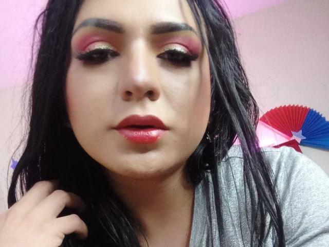 Iris Durand, met zwart haar, roze oogschaduw, gouden eyeliner, rode lippenstift en glanzende roze lippen, kijkt naar beneden met een lichtjes fronsende uitdrukking, en draagt een grijze blouse..