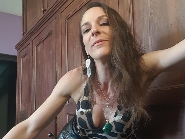 Natacha, com longos cabelos castanhos, vestindo uma blusa de estampa de leopardo com decote, um pingente verde e brincos grandes, apoiada em armários de madeira, sorrindo levemente..