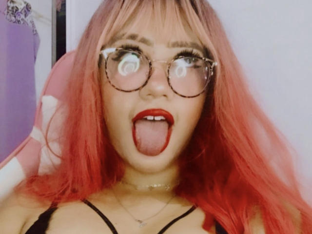 SexiestAngell com cabelo ruivo, óculos grandes e redondos, batom vermelho, língua de fora, com uma expressão engraçada, vestindo uma blusa preta com alças finas e um colar, sentada em uma cadeira rosa..