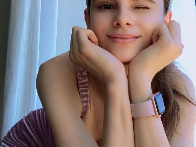 BatSama, les mains sur les joues, clignant de l'œil, portant une montre intelligente rose, vêtue d'un haut en velours violet, souriant doucement. Elle a une peau claire, des cheveux bruns attachés en queue de cheval, et des traits mignons..