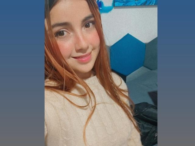 HeidyMillerr, con lunghi capelli castani, sorridente, indossa un maglione bianco in maglia, ha un piercing al naso e rossetto rosa, mostrando un viso carino e piccoli seni. Sfondo blu..