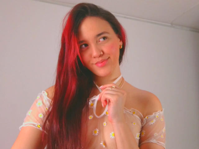 Milliee, con cabello largo de color rojo-negro, sonriendo, vestida con una blusa transparente con detalles en los hombros, sosteniendo su cuello, mostrando sus pechos, con un ligero rubor rosado en las mejillas..