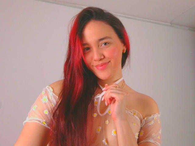 Milliee with red and black hair, smiling, wearing a sheer off-shoulder top with daisy flowers, holding a white ribbon, cute, small tits, light skin, pink lips.
