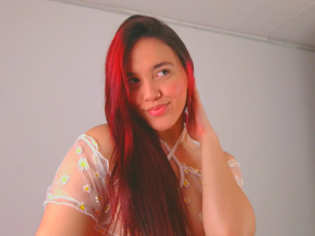 Milliee, met rood en zwart haar, draagt een doorschijnend topje met bloemen en raakt haar hoofd aan met een lichte glimlach, waarbij haar schattige, kleine borsten te zien zijn. Zachte, warme verlichting..