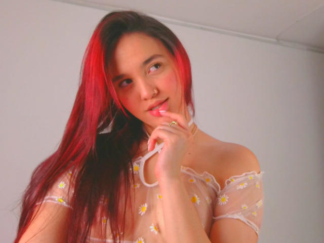 Milliee with red and black hair, wearing a sheer, off-shoulder top with daisy patterns, biting her finger, looking cute with pink blush on her cheeks.