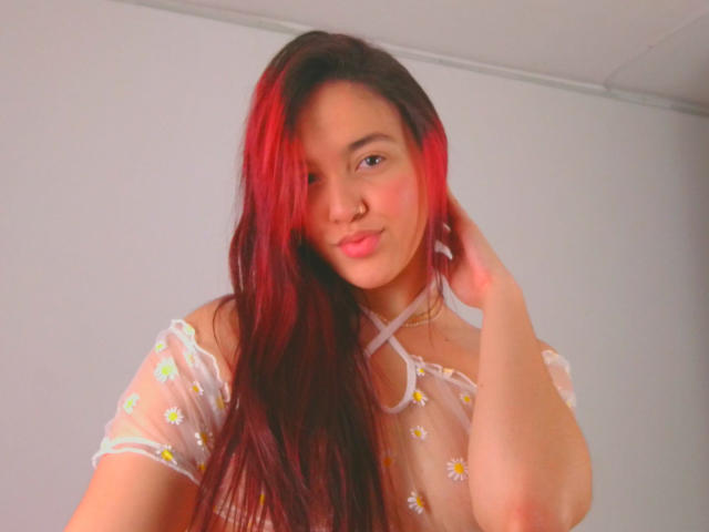 Milliee, com cabelo ruivo, vestindo uma blusa transparente com estampas de margaridas, tem a mão no cabelo, mostrando os seios e um piercing no nariz, sorrindo suavemente..