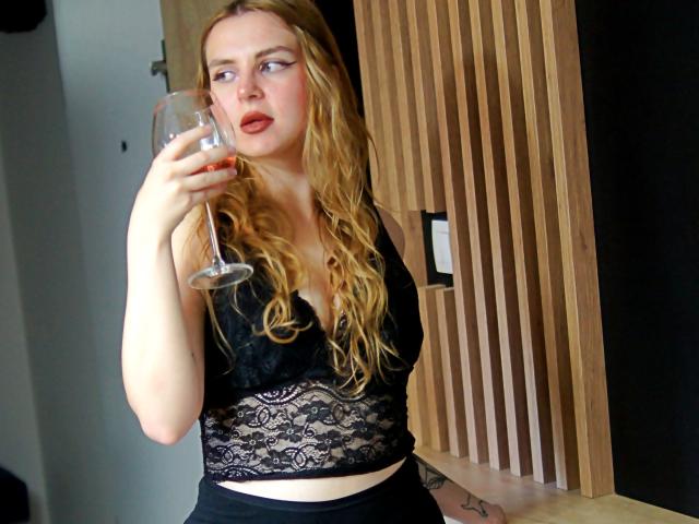 ZoeyFancy with long blonde hair, black lace top, and black pants, holding a glass of red wine, looking to the side with slightly parted red lips.