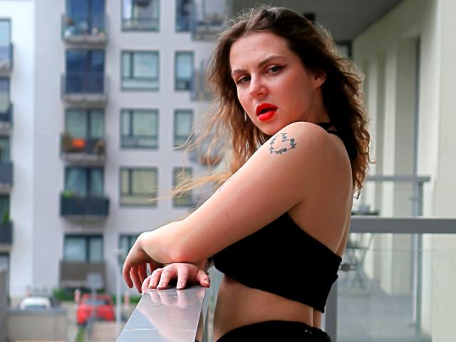 ZoeyFancy, avec ses cheveux bruns ondulés, son rouge à lèvres rouge, son haut noir, un tatouage sur l'épaule, appuyée contre une rambarde en métal, avec une expression légèrement agacée, dans un environnement urbain. Ses seins sont visibles, les bras croisés..