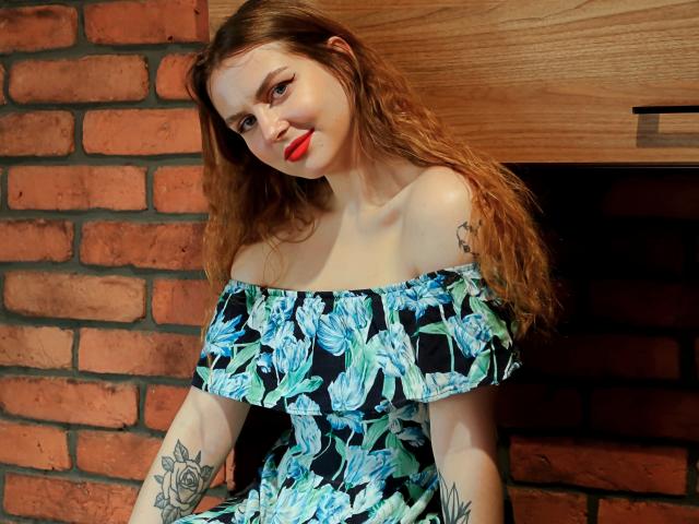 ZoeyFancy met rood haar, een blauwe bloemenjurk met open schouder, rode lippenstift, tatoeages op de armen, lachend, staand tegen een bakstenen muur, een houten kast op de achtergrond. Borsten, billen, borsten, vagina zijn niet zichtbaar..