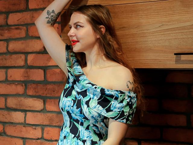 ZoeyFancy con rossetto rosso, abito floreale blu-verde, a spalle scoperte, braccio alzato, mostrando un tatuaggio, capelli castani ondulati, mobiletto in legno, parete di mattoni, sorridente, carina, con il seno parzialmente visibile..