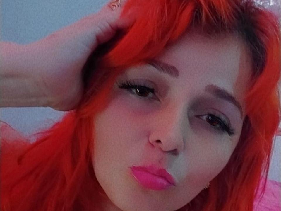 KyleGinger69 met felrood haar, hand in het haar, volgezwollen roze lippen, dikke zwarte eyeliner en een lichte blos, kijkt met een verleidelijke uitdrukking in de camera..