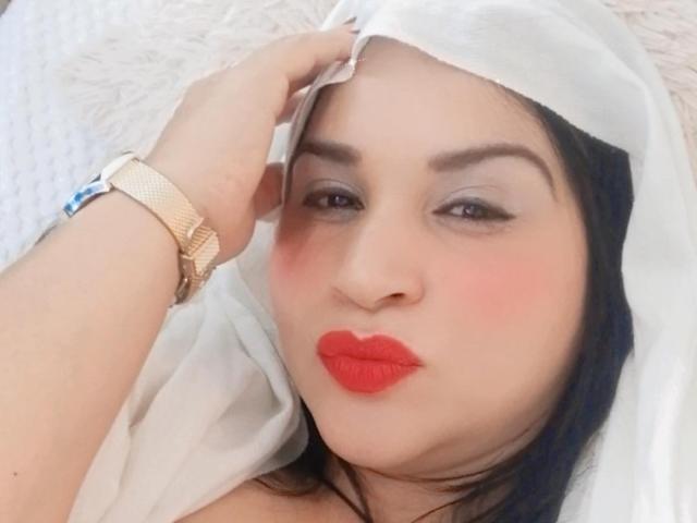 JameelaX with red lipstick, flushed cheeks, and a white sheer head covering, touching her forehead, wearing a gold watch, has dark hair, and light skin, looking at the camera.