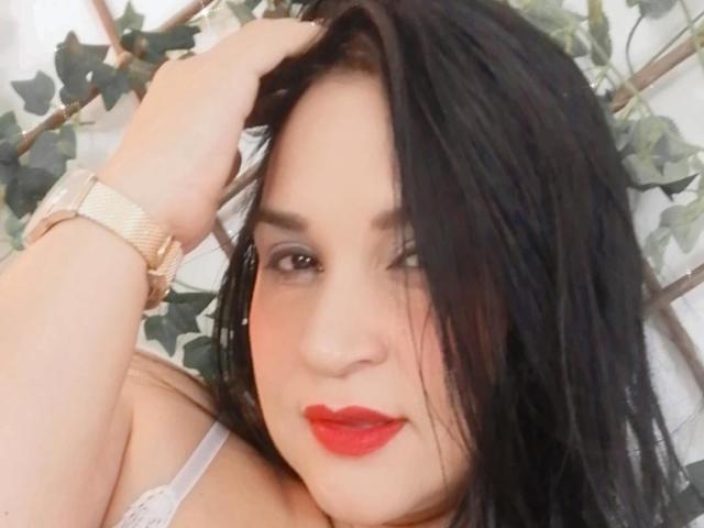 JameelaX with black hair, red lipstick, wearing a white bra, gold watch, hand in hair, looking at camera, slightly messy hair, soft smile, fair skin, cute face.