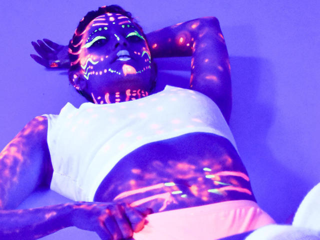 JaneRoa with dark purple, neon-lit skin, green eyes, white polka-dot tank top, pink shorts, laying back, arms behind head, glowing patterns on face and body, relaxed.