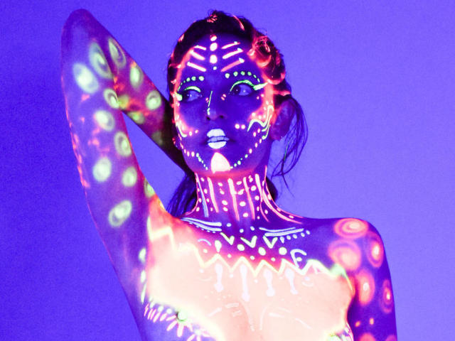 JaneRoa's glowing, neon-patterned body is lit up with colorful, glowing spots and lines on her face, chest, and arm, raising her hand against a purple backdrop.
