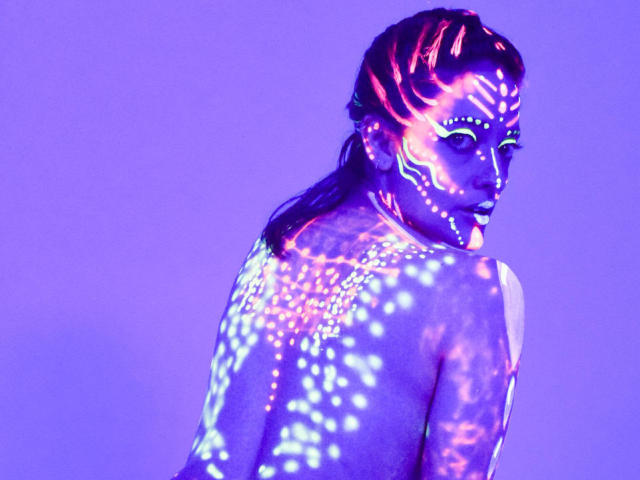 JaneRoa's glowing purple and neon blue body paint, with bright patterns on her back and face, looks sexy and illuminated. She's topless, turning slightly to look at the camera.