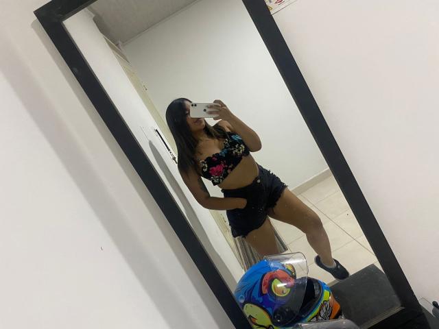 Valery, les cheveux foncés, portant un haut sans bretelles à motifs floraux et un short en jean noir, prend un selfie devant un miroir dans une pièce blanche. Ses seins sont visibles, et elle tient son téléphone..