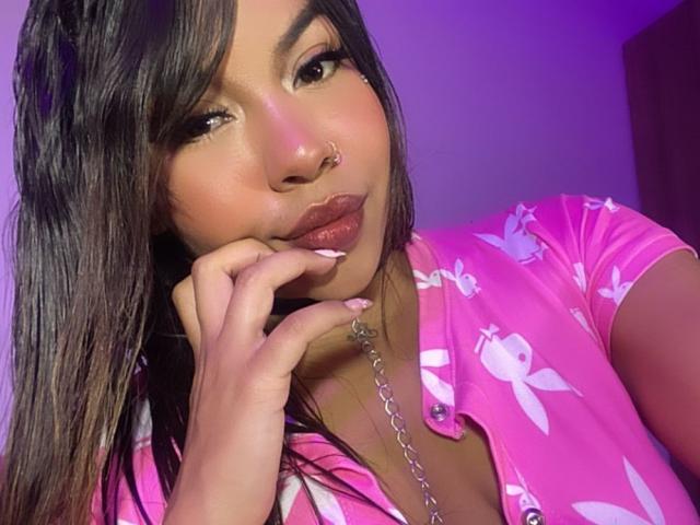 Valery: Cabelos pretos e lisos, olhos castanhos, piercing no nariz e lábios rosados brilhantes, vestindo uma camisa rosa com estampas florais, tocando o queixo com um colar de prata..