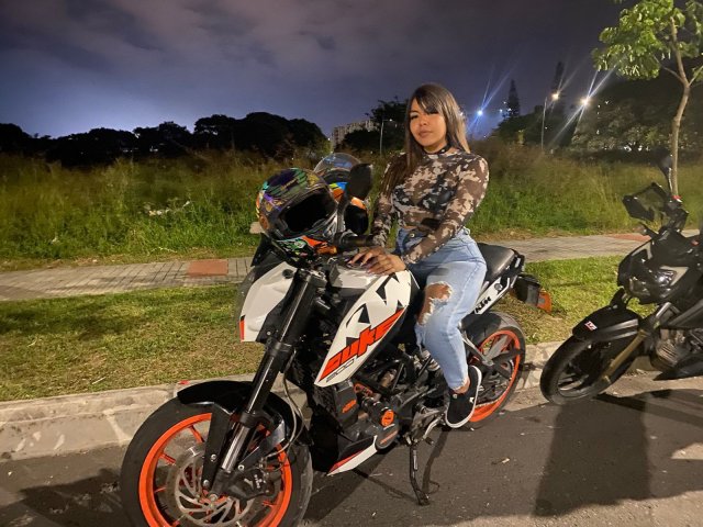 Valery, die aan het spuiten is met haar achterwerk op een zwart-wit motorfiets, draagt een camouflagehemd en gescheurde jeans, en staat 's nachts met een zwarte motorfiets op de achtergrond..