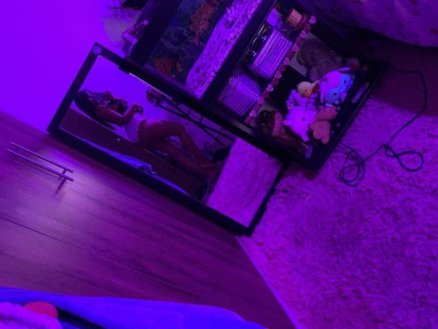O reflexo em um espelho mostra o traseiro e os seios nus de ValerySquirting, deitada em uma cama com iluminação roxa, cercada por brinquedos de pelúcia em um chão coberto de carpete..