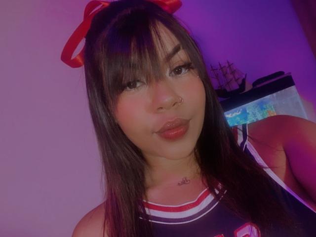 ValerySquirting tiene el pelo largo y negro, un lazo rojo, un piercing en la nariz y lleva una camiseta de animadora en blanco y negro. Sus pechos son pequeños y está haciendo pucheros con labios brillantes..