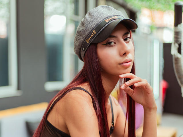 DalilaRusso with long red hair, wearing a gray cap and black strap top, looking over her shoulder with a subtle smile, fingers near her lips, showing her tits.