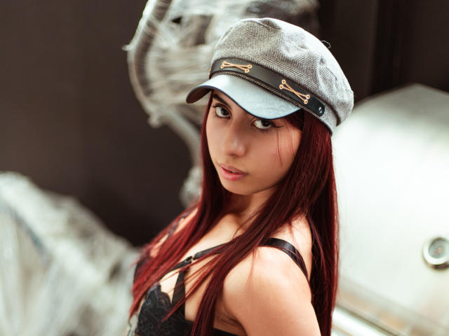 DalilaRusso with red hair, wearing a gray cap and black lace bra, looking over her shoulder, showing her tits, in a softly lit, indoor setting.