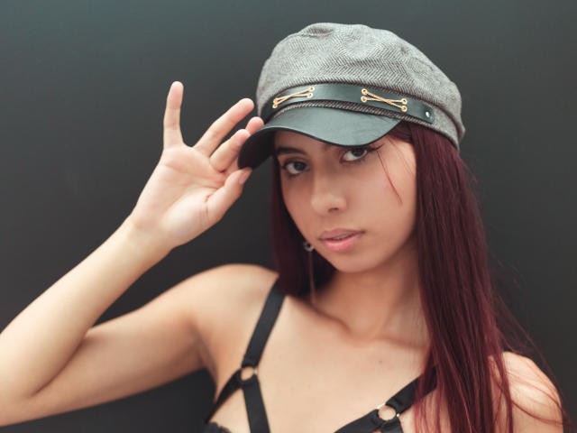 DalilaRusso with red hair, wearing a grey cap with gold buttons, black strappy top, and looking at the camera, hand on cap, showing off her tits.