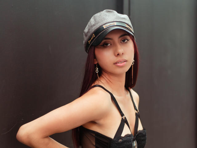 DalilaRusso with long red hair, wearing a gray cap, black lace bra, and gold earrings, has her hand on her hip, showing her tits and a slight smile.
