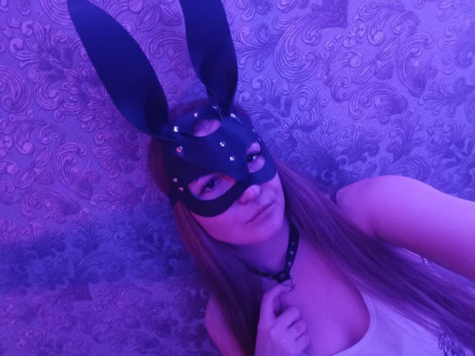 CanndyDi, avec de longs cheveux bruns, portant un masque noir avec des paillettes violettes et des oreilles de lapin noires, un débardeur blanc, un éclairage violet et un collier noir, avec une expression légèrement boudeuse..