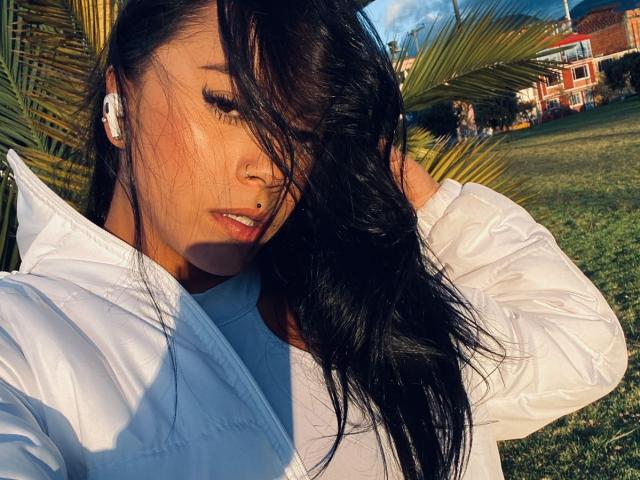 Amberv with black hair, white jacket, and white earbuds, eyes closed, sunlight in face, hands in hair, nose ring, and a grassy background with palm leaves.