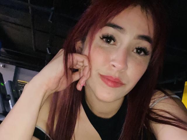 NicoColeman with red hair, brown eyes, and pink lipstick, touching her cheek with a hand, smiling slightly, wearing a black halter top, in a dimly lit room.