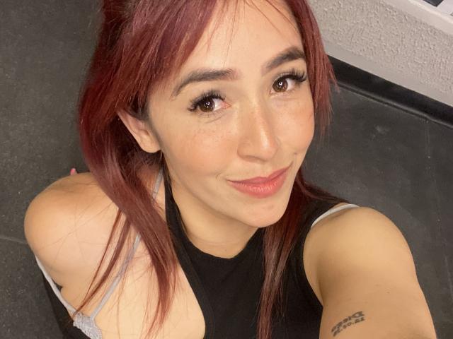 NicoColeman with red hair, brown eyes, and freckles, smiling at the camera, showing her tits in a black sleeveless top, tattoo on her right arm, selfie.