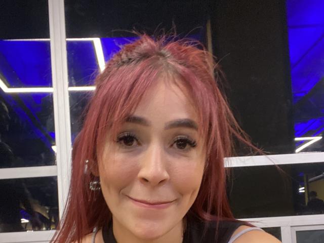 NicoColeman has red hair, brown eyes, and a cute smile. She's wearing a black top with small earrings. Her tits are small, and she looks sexy.