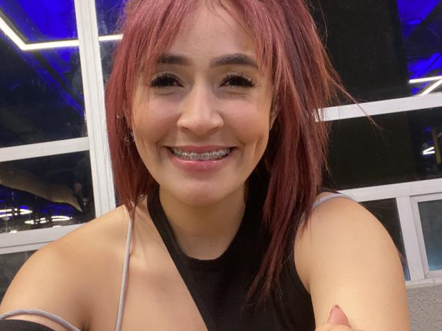 NicoColeman has red hair, smiling with metal teeth, wearing a black top, and showing her tits slightly in a selfie. She's cute and has a sexy smile.