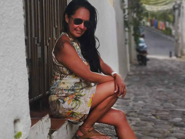 LailaCastillo, met donker haar, gekleed in een bloemenjurk, zwarte zonnebril, bruine sandalen, hurkt op een geplaveide pad, met een witte muur en houten latten op de achtergrond, lachend, met een tatoeage op de rechter bovenarm, bruine huid..